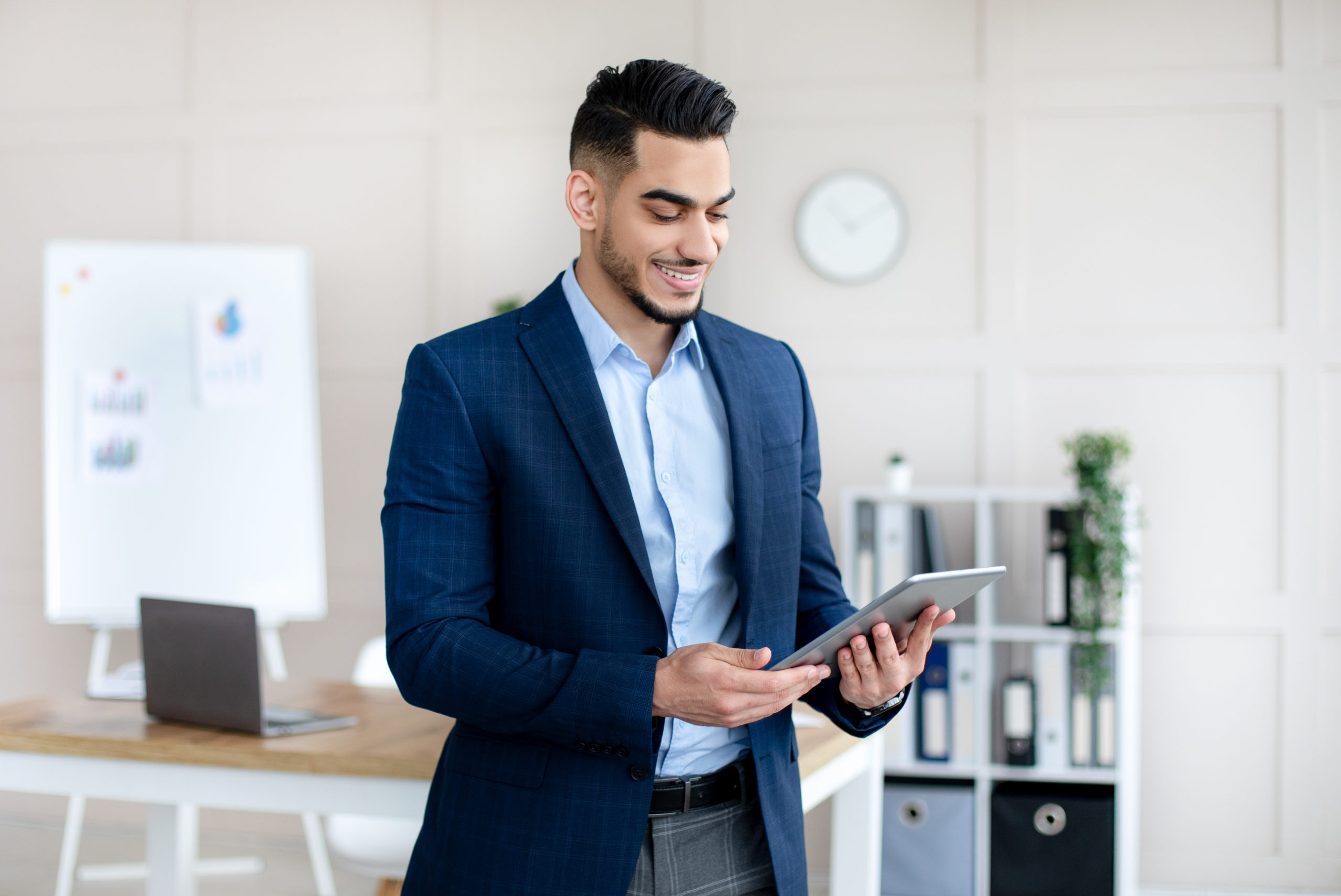 Portrait of young Arab man in formal suit working online, using digital tablet in modern office. Millennial Eastern guy reading documents, checking email or browsing web for information on touch pad