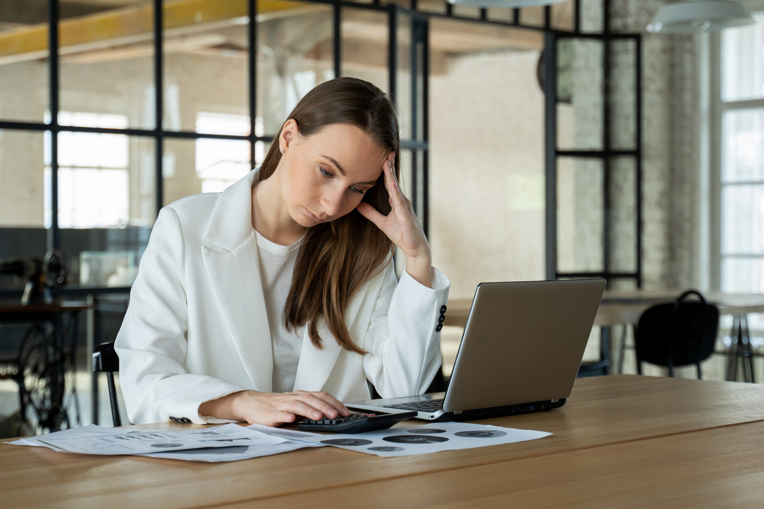 Stylish young female accountant looking through finances, using a laptop and a calculator, experiencing stress due to the fact that the numbers do not add up in the report.